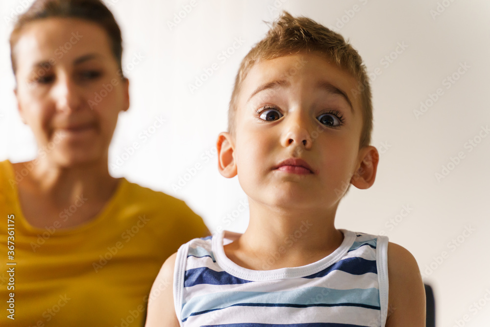 Front view on small caucasian boy making face looking serious stiff while sitting by his mother at home in day Front view on small caucasian boy making face looking serious stiff while sitting by his mother at home in day