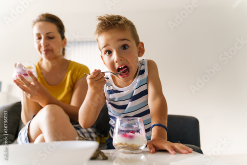 Front view on small boy eat desert by the table while his mother is watching in background at home - little child having cake hold spoon while eating in summer day - real people childhood concept