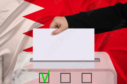 female voter drops a ballot in a transparent ballot box against the backdrop of the Bahrain national flag, concept of state elections, referendum