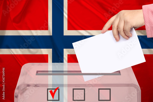 female voter lowers the ballot in a transparent ballot box against the background of the national flag of Norway, concept of state elections, referendum