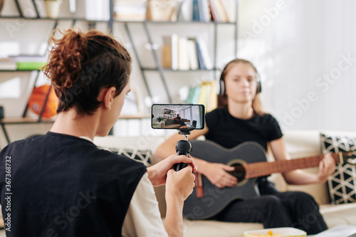 Teenage boy using smartphone when shooting his friend playing guitar and singing for some contest