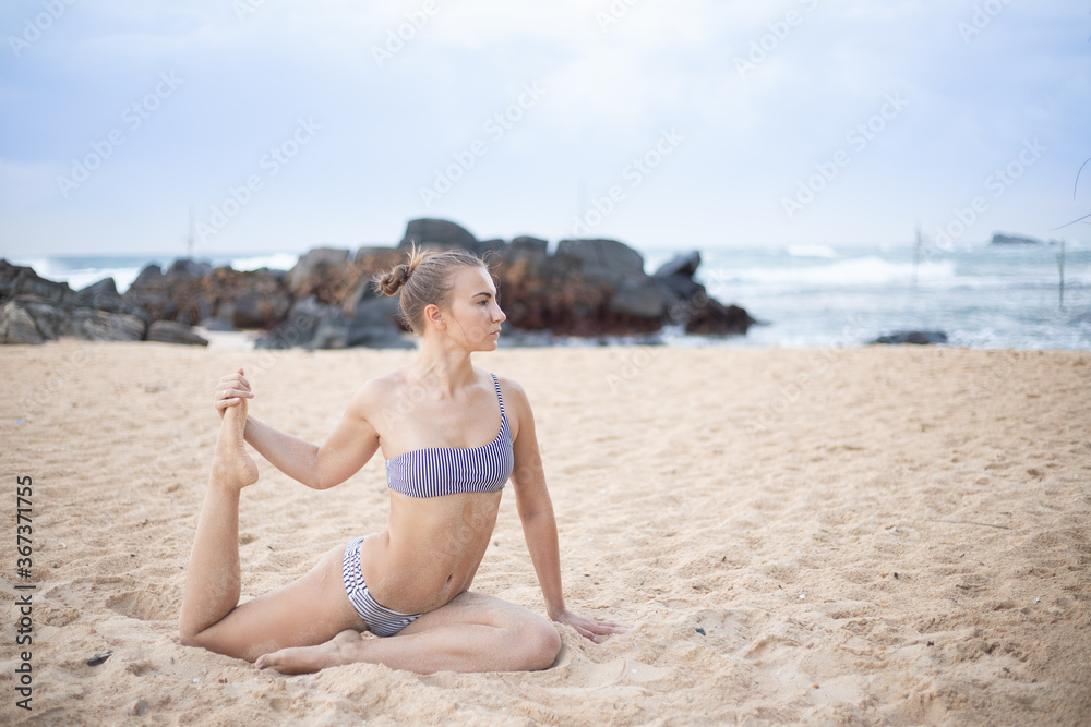 Young attractive girl in a swimsuit practices yoga on the beach against a blue sky