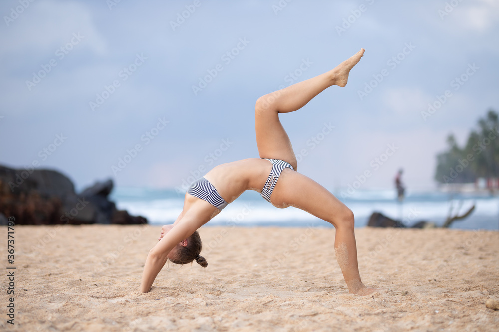 Young attractive girl in a swimsuit practices yoga on the beach against a blue sky Young attractive girl in a swimsuit practices yoga on the beach against a blue sky