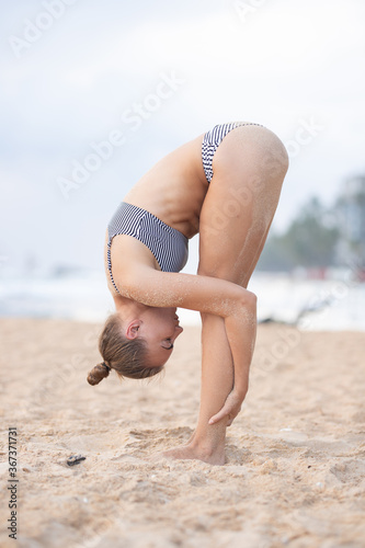Young attractive girl in a swimsuit practices yoga on the beach against a blue sky