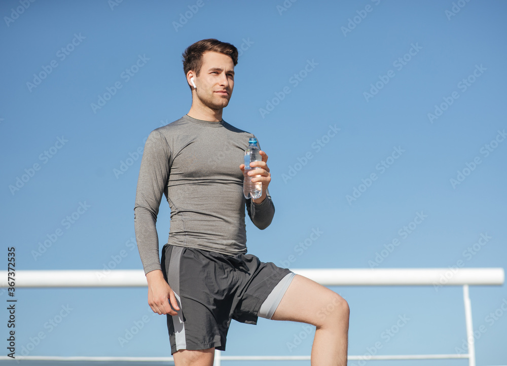 Fototapeta premium Health and body care. Muscular handsome guy in sportswear, with wireless headphones holding bottle of water on blue sky background