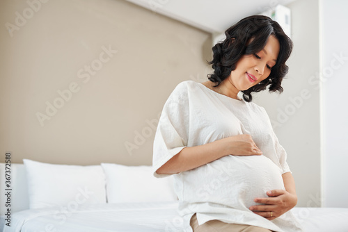Happy pregnant young Asian woman sitting on bed and touching her belly