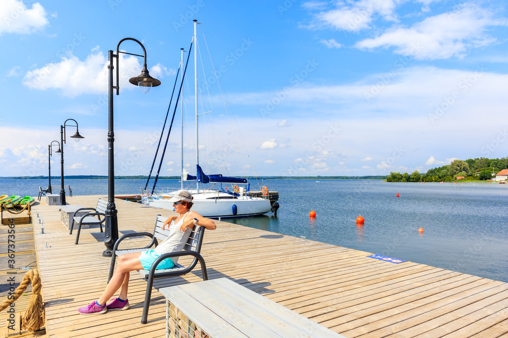 Young woman sitting on pier and looking at boats anchoring in  sailing port Nowe Guty on Lake Sniardwy on summer sunny day,  Masurian Lakes, Poland