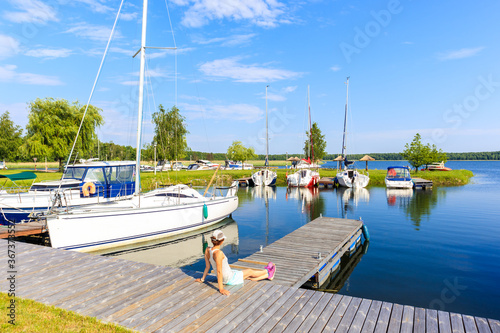 Young woman sitting on pier and looking at boats anchoring in Pisz sailing po...