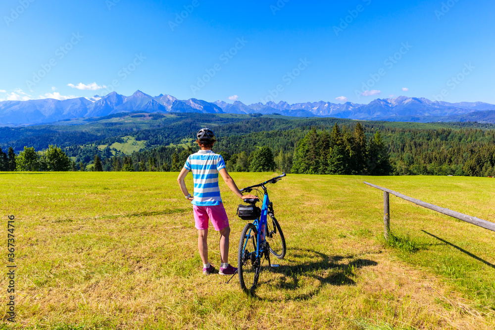Fototapeta premium Young female biker standing with bicycle on meadow and looking at panorama of Tatra Mountains on beautiful summer sunny day at Lapszanka Pass, Poland