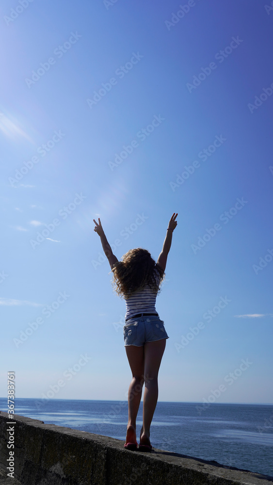 A slender curly-haired girl in denim shorts on the background of the blue sea on a sunny day, summer vacation. model shows V sign, world at peace A slender curly-haired girl in denim shorts on the background of the blue sea on a sunny day, summer vacation. model shows V sign, world at peace
