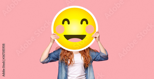Redhead girl hiding her face behind happy emoji smile