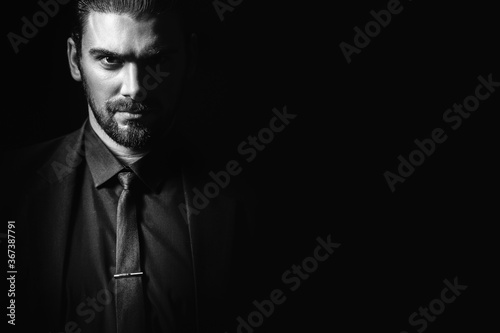 Portrait of a white Caucasian male brunet in a black classic suit, black shirt and black tie over a black background. Fashionable portrait of an attractive man with dark hair.