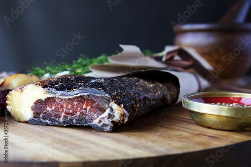 A piece of dried meat biltong on a wooden board wrapped in brown paper.  A small bowl of coriander seed sits to the side and a pestal and mortar against a black background  photo