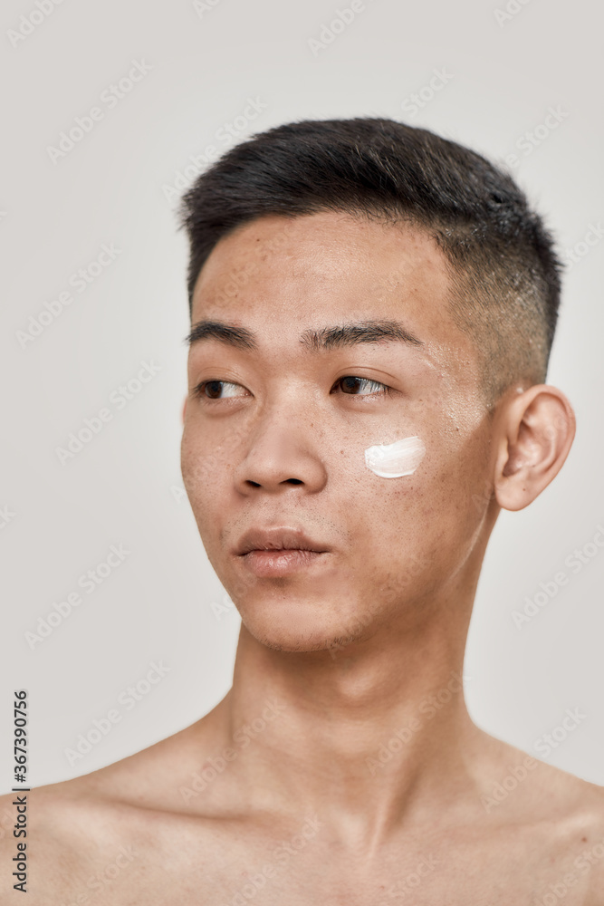 Obraz premium Perfect skincare. Close up portrait of shirtless young asian man with cream applied on his cheek looking aside isolated over white background. Beauty, skincare routine concept