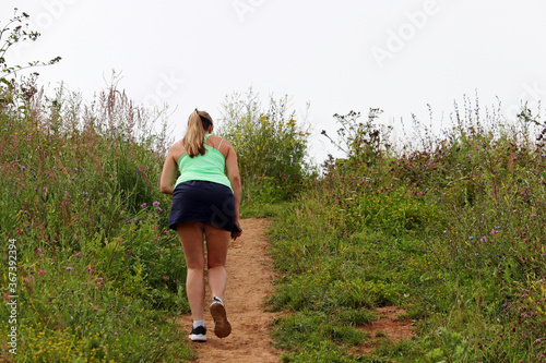 Girl in short skirt climbs to the top on a path, rear view. Summer meadow overgrown of grass and wildflowers on sky background, hiking and slimming concept