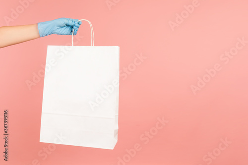 Profile side view closeup of human hand in blue surgical gloves holding and showing white shopping bag. indoor, studio shot, isolated on pink background.