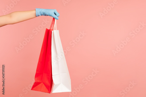 Profile side view closeup of human hand in blue surgical gloves holding and showing red and white shopping bag. indoor, studio shot, isolated on pink background.