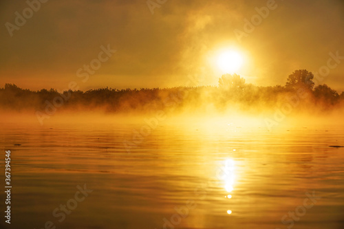 A fiery sunrise is reflected in the calm waters, over trees in the bright orange sky, mist and light waves on the water surface