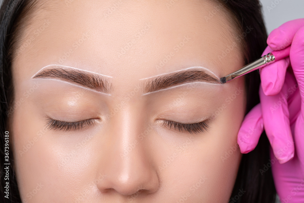 Brunette teenager girl having permanent makeup tattoo on her eyebrows.  Make-up artist makes markings with white paste for eyebrow tattooing.  Professional makeup and skin care cosmetology. Photos | Adobe Stock