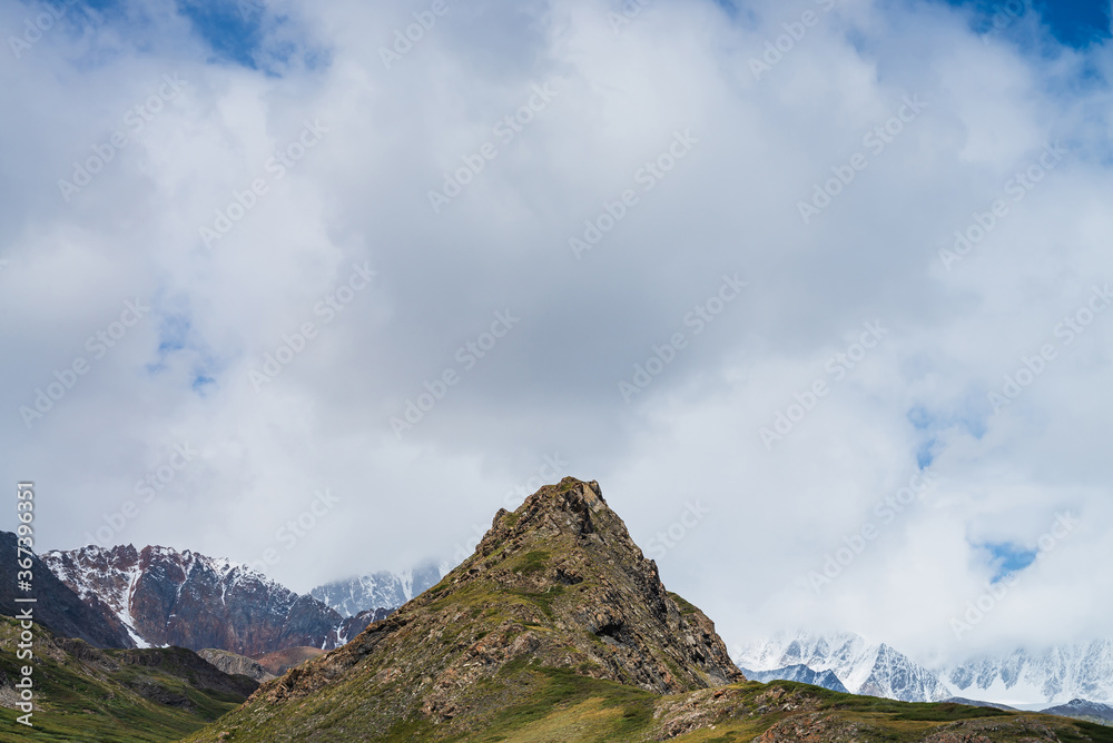 Atmospheric alpine landscape with beautiful pinnacle rock and low clouds in green valley among snowy mountains. Awesome view to sharp top of green hill and mountain range with snow under cloudy sky.