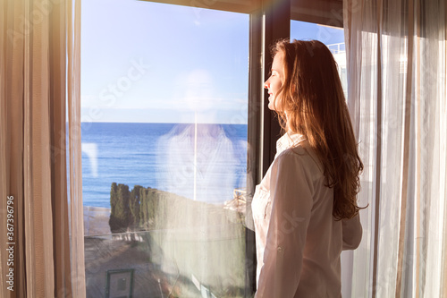 Beautiful long-haired woman in a white shirt standing indoor by a panoramic window with curtains with view on the sea and enjoying the morning sun lights with her eyes closed.