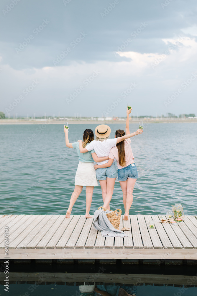 three girls stand on a wooden pier in front of the water. rest, picnic,lemonade. back view
