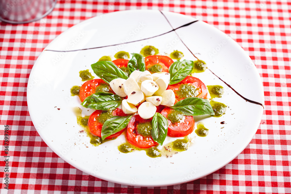 Traditional Italian caprese salad with sliced tomatoes, mozzarella cheese, basil and pesto sauce on white plate. Close up
