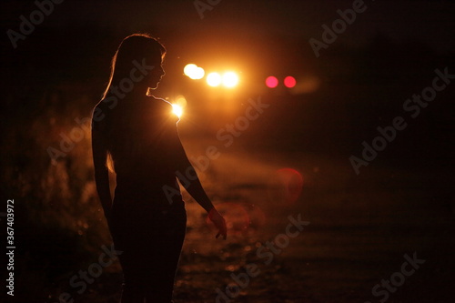 Silhouette of young slender woman in the backlight of car headlights on the road