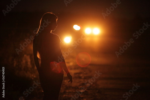 Silhouette of young slender woman in the backlight of car headlights on the road