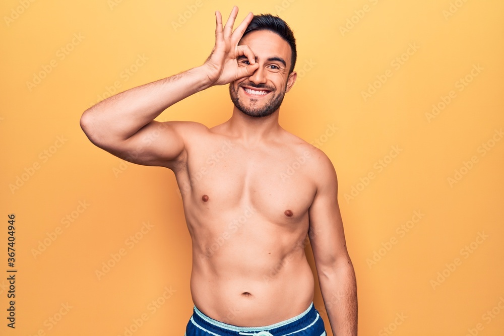 Young handsome man with beard wearing sleeveless t-shirt standing over yellow background smiling happy doing ok sign with hand on eye looking through fingers