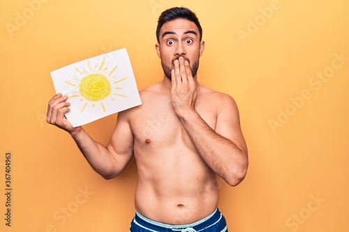 Young handsome man with beard on vacation wearing swimwear holding paper with sun draw covering mouth with hand, shocked and afraid for mistake. Surprised expression