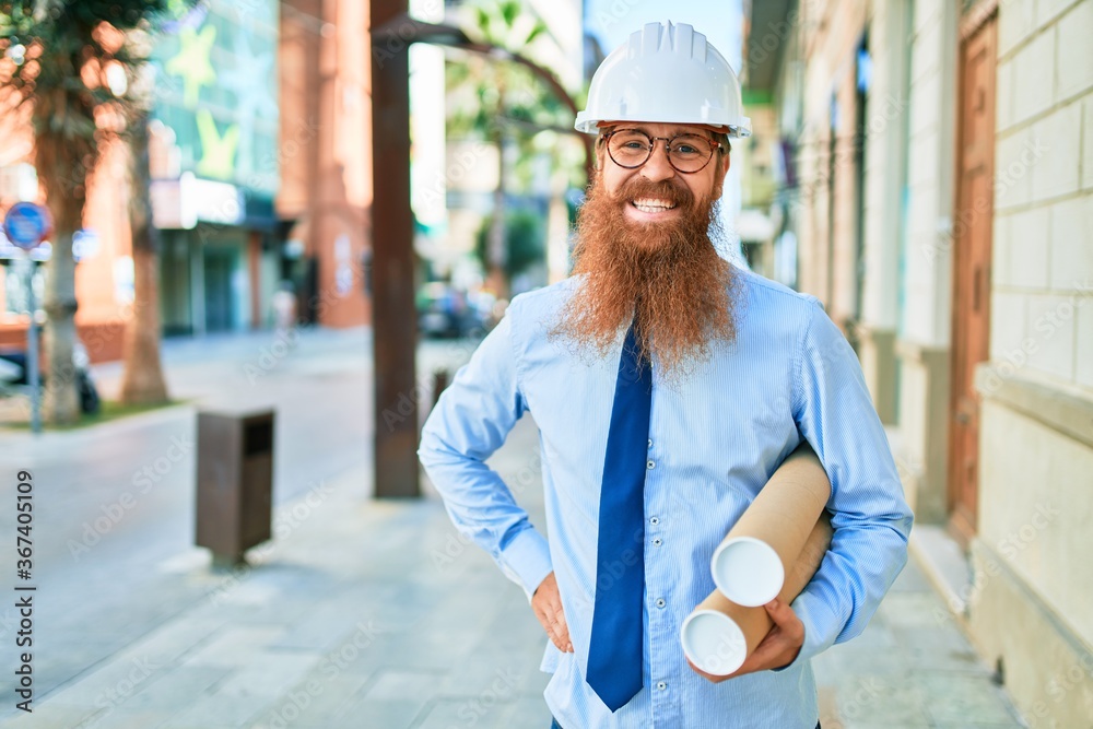 Young redhead architect man with long beard wearing hardhat smiling happy. Standing with smile on face holding blueprints at street of city.