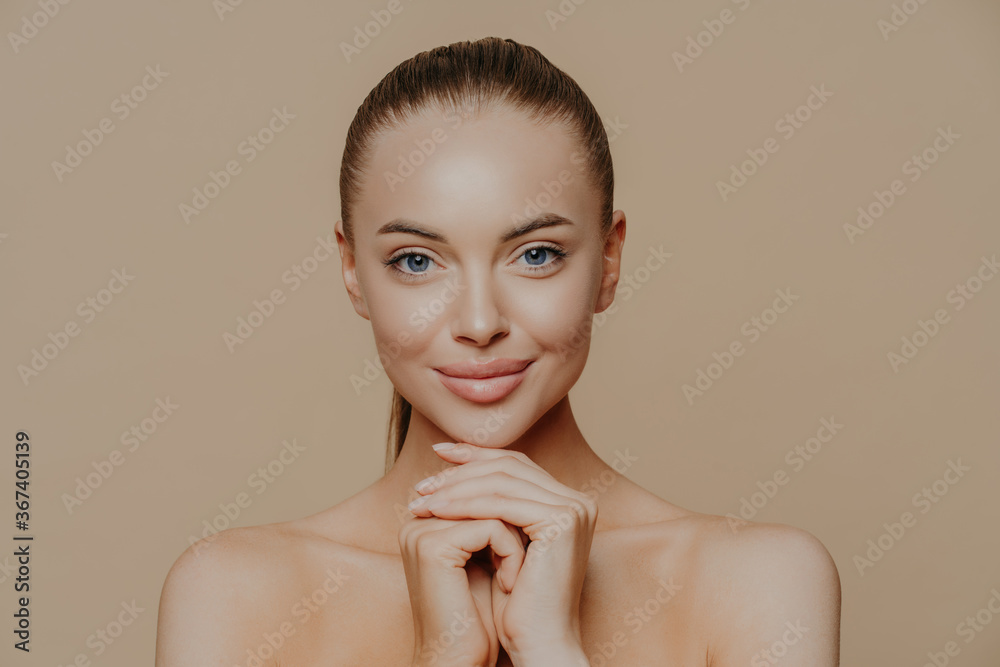 Young pretty woman with beauty skin and combed hair, keeps hands under chin, has healthy complexion after face purification, soft well cared body, isolated over studio wall. Facial treatment Young pretty woman with beauty skin and combed hair, keeps hands under chin, has healthy complexion after face purification, soft well cared body, isolated over studio wall. Facial treatment