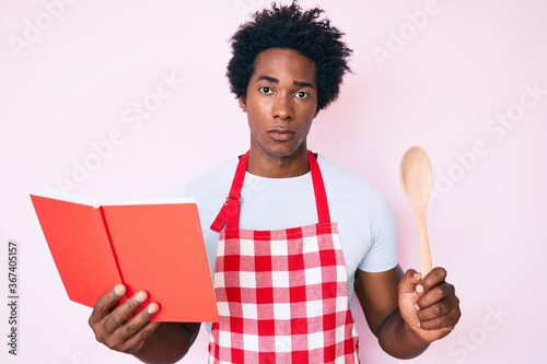 Handsome african american man with afro hair wearing professional baker apron reading cooking recipe book skeptic and nervous, frowning upset because of problem. negative person.