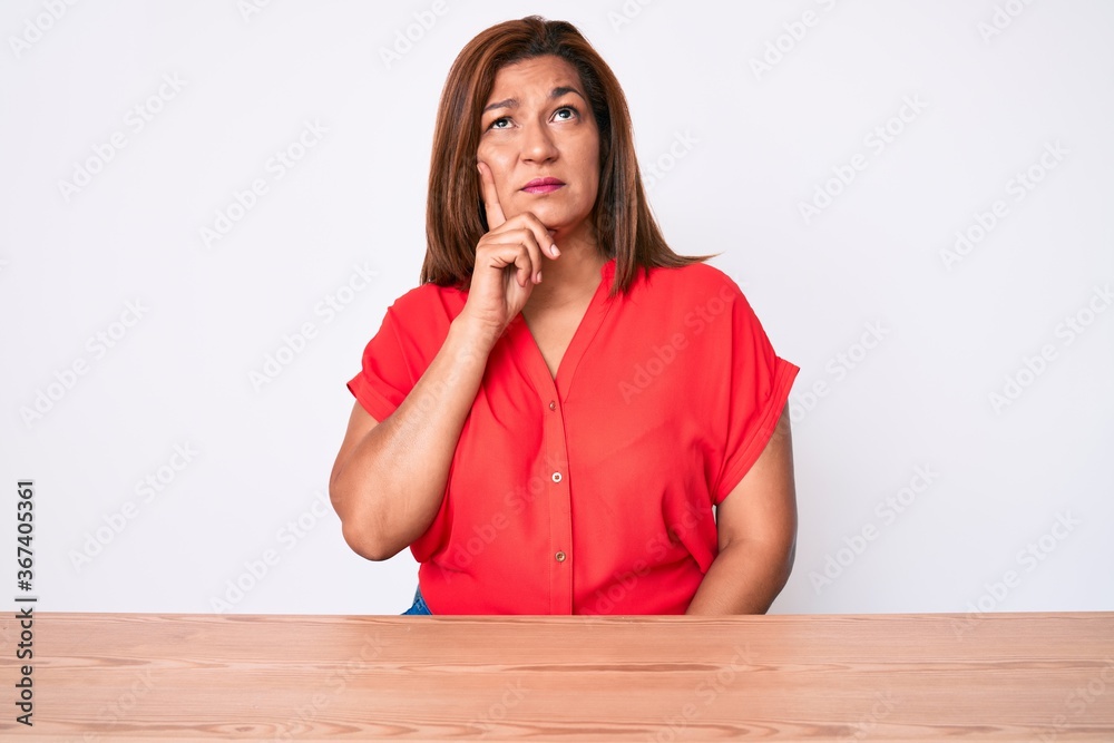 Middle age brunette hispanic woman wearing casual clothes sitting on the table thinking concentrated about doubt with finger on chin and looking up wondering Middle age brunette hispanic woman wearing casual clothes sitting on the table thinking concentrated about doubt with finger on chin and looking up wondering