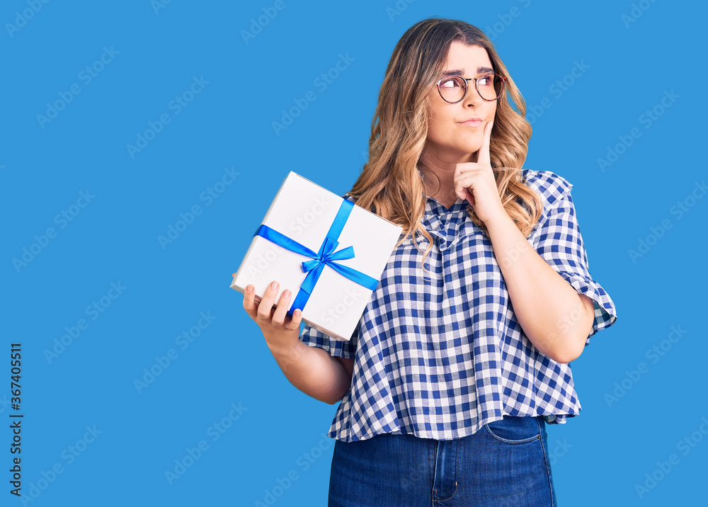 Young caucasian woman holding gift serious face thinking about question with hand on chin, thoughtful about confusing idea Young caucasian woman holding gift serious face thinking about question with hand on chin, thoughtful about confusing idea
