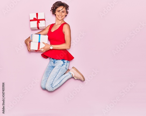Young beautiful woman holding birthday gift smiling happy. Jumping with smile on face over isolated pink background