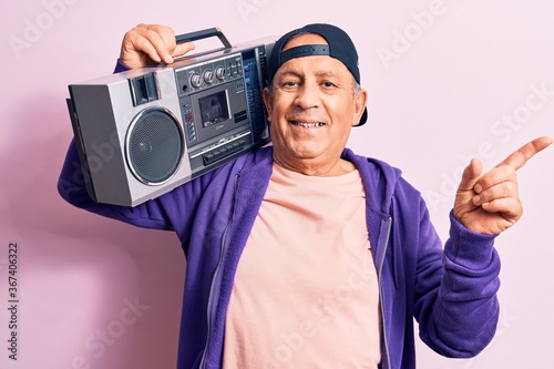 Senior handsome grey-haired modern man listening to music using vintage boombox smiling happy pointing with hand and finger to the side