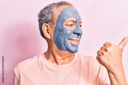 Senior man with grey hair wearing mud mask pointing thumb up to the side smiling happy with open mouth