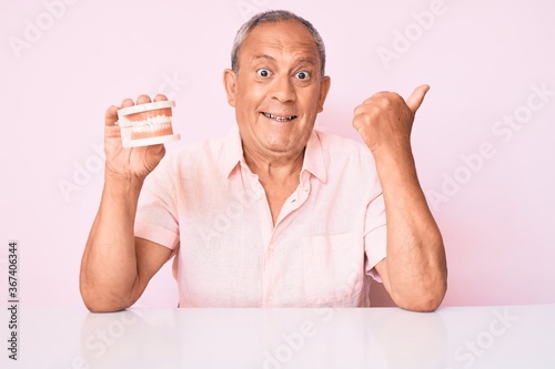 Senior handsome man with gray hair holding denture sitting on the talbe pointing thumb up to the side smiling happy with open mouth photo