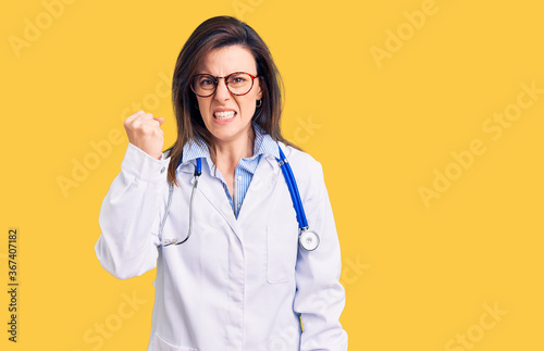 Young beautiful woman wearing doctor stethoscope and glasses angry and mad raising fist frustrated and furious while shouting with anger. rage and aggressive concept.