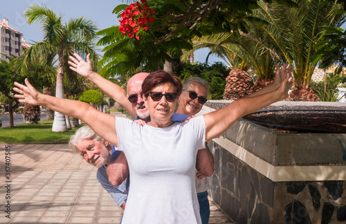 Cheerful group of senior friends enjoying vacation - four retired people in public park with palms trees and blossom plants - active retirement concept