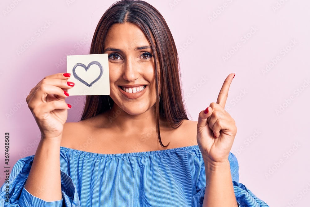 Young beautiful brunette woman holding reminder with heart smiling happy pointing with hand and finger to the side Young beautiful brunette woman holding reminder with heart smiling happy pointing with hand and finger to the side