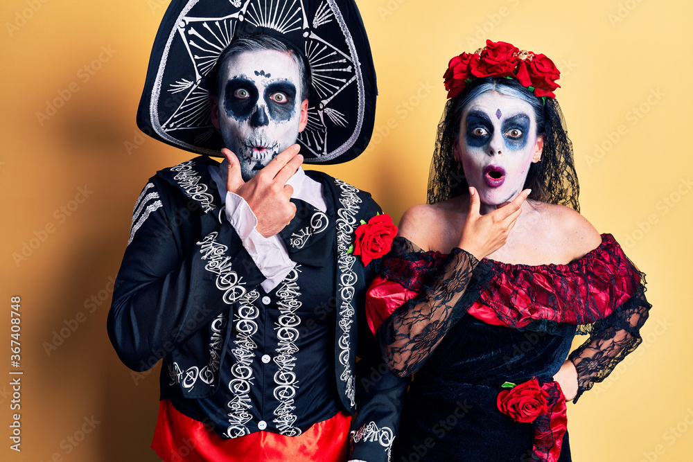 Young couple wearing mexican day of the dead costume over yellow looking fascinated with disbelief, surprise and amazed expression with hands on chin Young couple wearing mexican day of the dead costume over yellow looking fascinated with disbelief, surprise and amazed expression with hands on chin