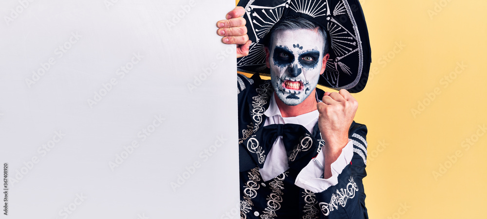 Young man wearing mexican day of the dead costume holding blank empty banner annoyed and frustrated shouting with anger, yelling crazy with anger and hand raised