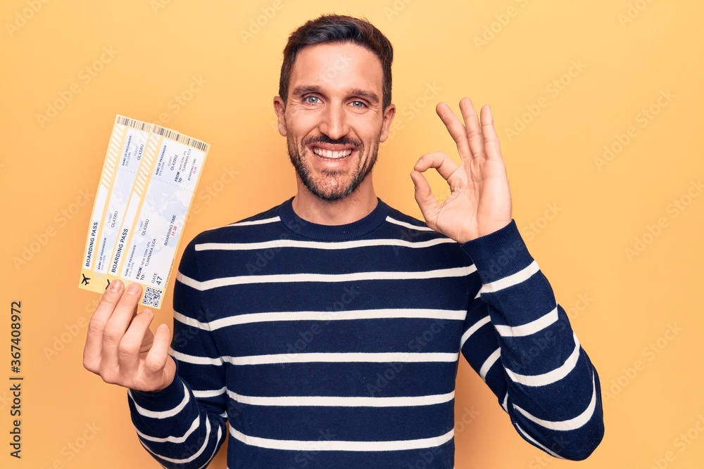 Young handsome tourist man on vacation holding airline boarding pass over yellow background doing ok sign with fingers, smiling friendly gesturing excellent symbol Young handsome tourist man on vacation holding airline boarding pass over yellow background doing ok sign with fingers, smiling friendly gesturing excellent symbol