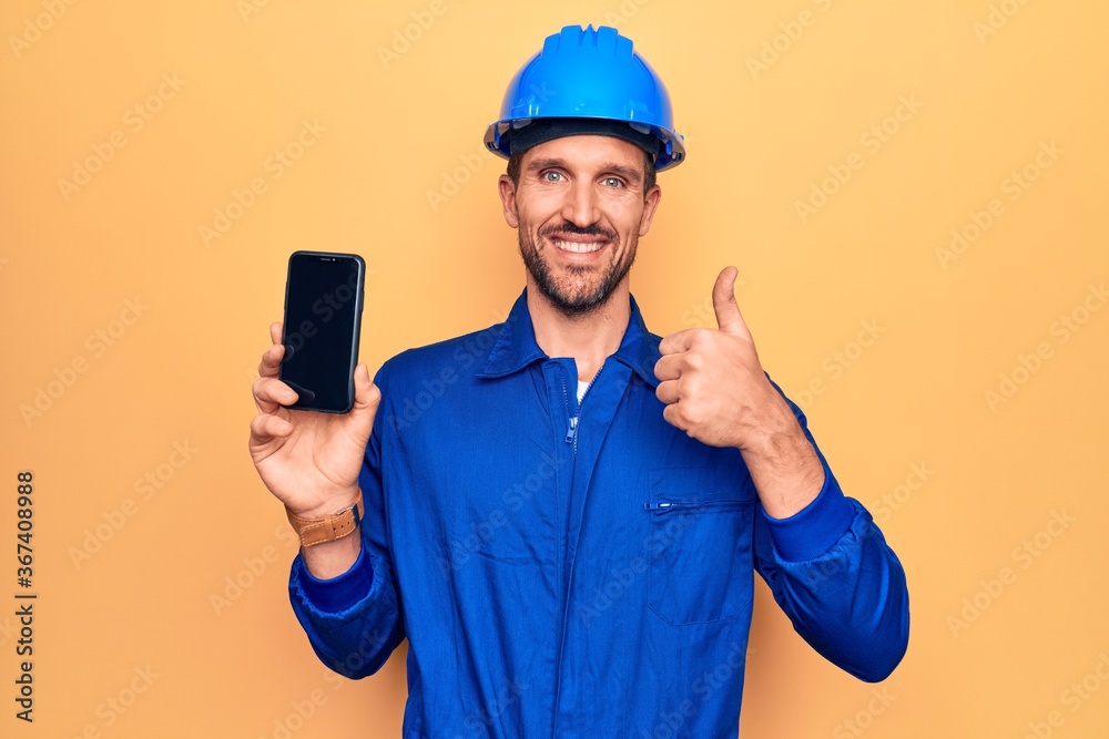 Young handsome worker man wearing uniform and hardhat holding smartphone showing screen smiling happy and positive, thumb up doing excellent and approval sign