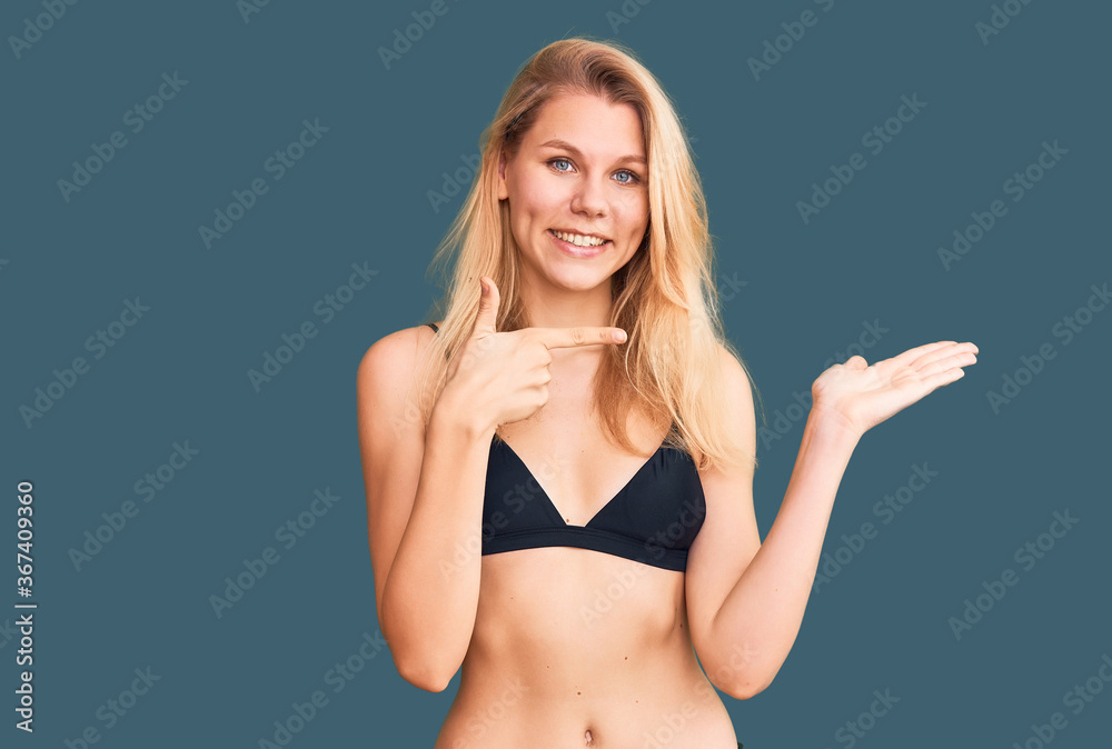 Fototapeta premium Young beautiful blonde woman wearing bikini amazed and smiling to the camera while presenting with hand and pointing with finger.