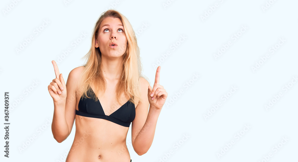 Young beautiful blonde woman wearing bikini amazed and surprised looking up and pointing with fingers and raised arms. Young beautiful blonde woman wearing bikini amazed and surprised looking up and pointing with fingers and raised arms.
