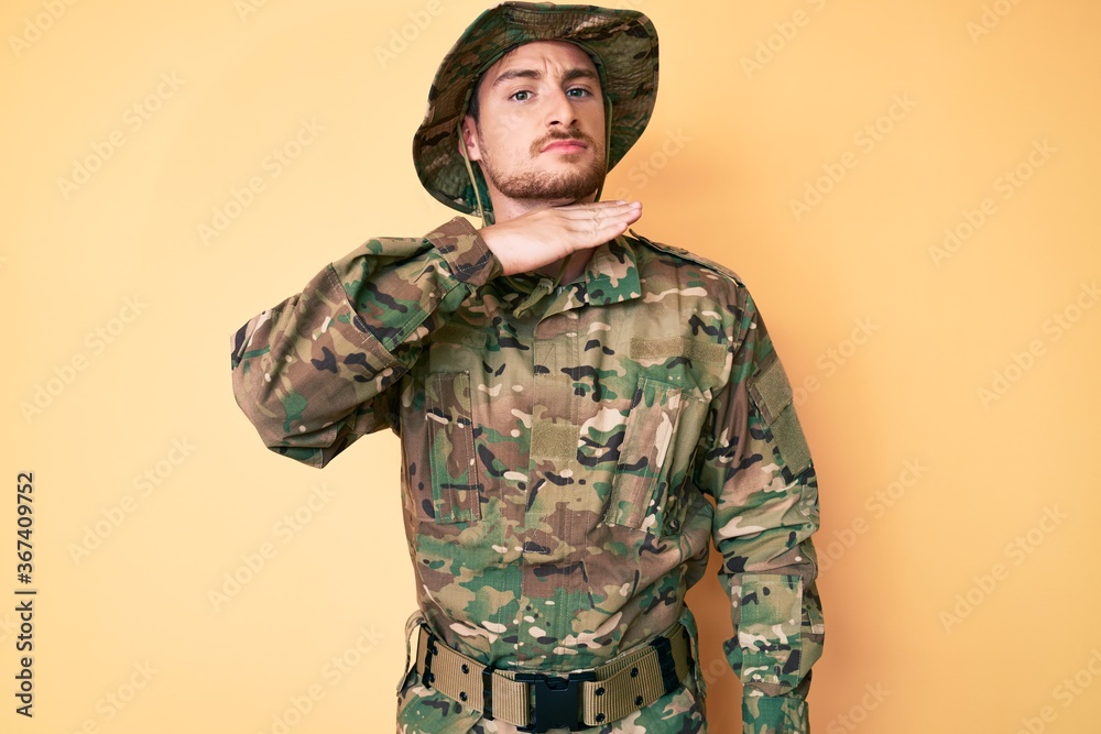 Young caucasian man wearing camouflage army uniform cutting throat with hand as knife, threaten aggression with furious violence
